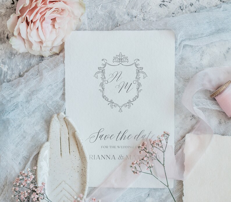 K&ouml;nnte beinhalten: Save-the-Date-Karte mit einem zarten floralen Design und einem Monogramm der Buchstaben "R" und "M" in einem dekorativen Rahmen. Der Text lautet "Save the date for the wedding of Rianna & Matthew".
