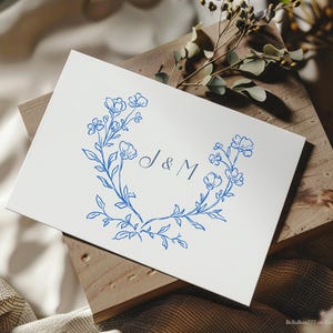 May include: A white card with a blue floral design and the initials "J & M" in the center. The floral design is a wreath of small blue flowers and leaves.