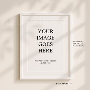 May include: White frame mockup with the text "YOUR IMAGE GOES HERE" and "FITS YOUR DIN A RATIO IMAGE HERE". The text "PSD WITH SMART OBJECT & JPEG FILE" is also included. The frame is on a beige wall with shadows.