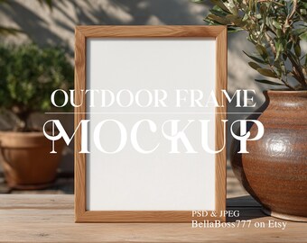 May include: A wooden picture frame mockup with a white interior, set outdoors on a wooden surface. The frame is surrounded by greenery, including a potted plant and a decorative vase. The text "OUTDOOR FRAME MOCKUP" is displayed on the frame.