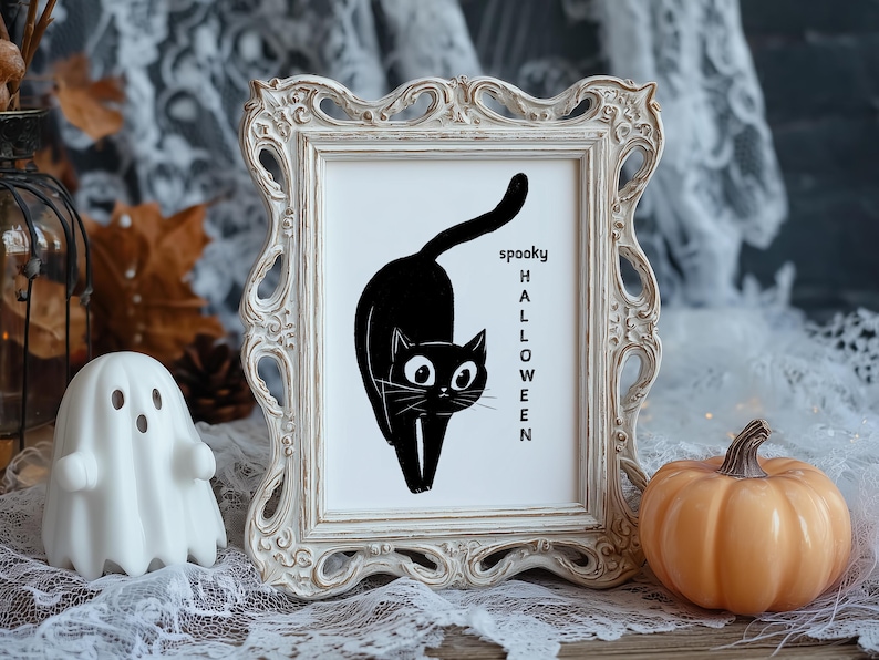 May include: A Halloween-themed print in a decorative white frame, with a black cat illustration and the words "spooky HALLOWEEN". An orange pumpkin and a white ghost ornament are placed nearby. The scene is set on a wooden surface with lace.