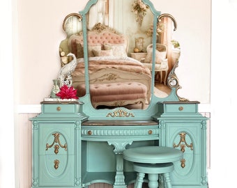 May include: A vintage vanity set with a large, ornate mirror, two side cabinets, and a stool. The vanity is painted a light teal blue with gold accents. The mirror has a gold frame with intricate carvings. The vanity has a marble top and the stool has a round seat and four legs.