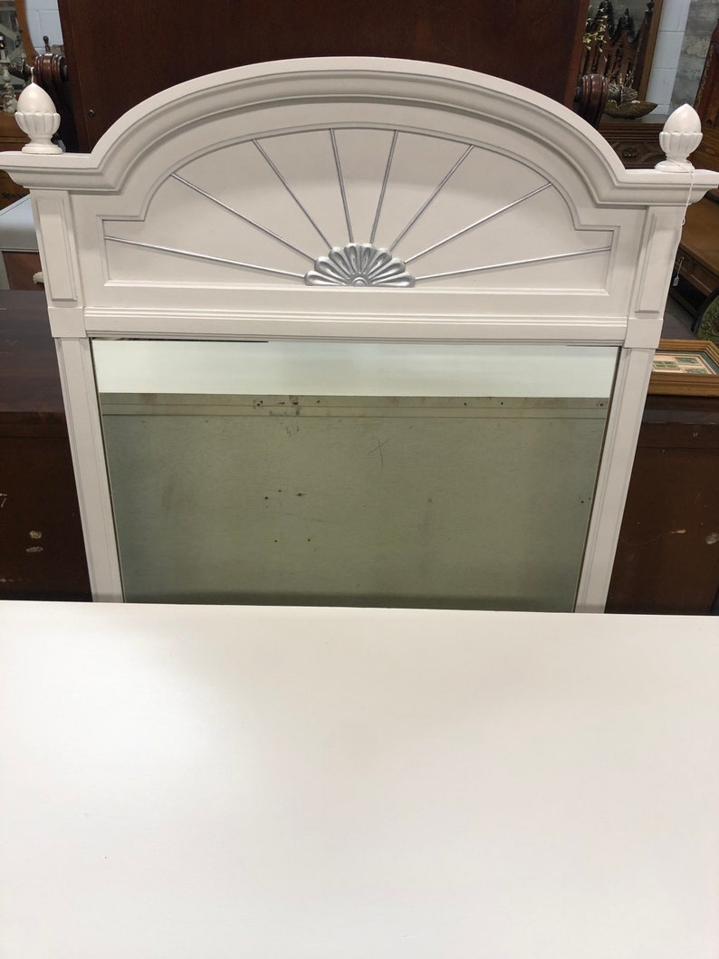 Pu&ograve; includere: Specchio con cornice in legno bianco con un design decorativo a raggiera argentato e uno specchio bisellato.