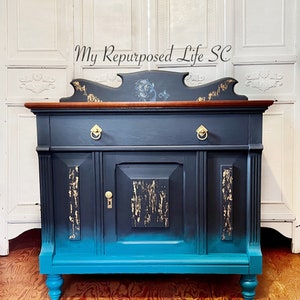 May include: A dark blue and teal painted antique sideboard with gold accents. The top of the sideboard has a carved wooden design and a wooden top. The sideboard has two doors and two drawers. The sideboard is sitting on a wooden floor.