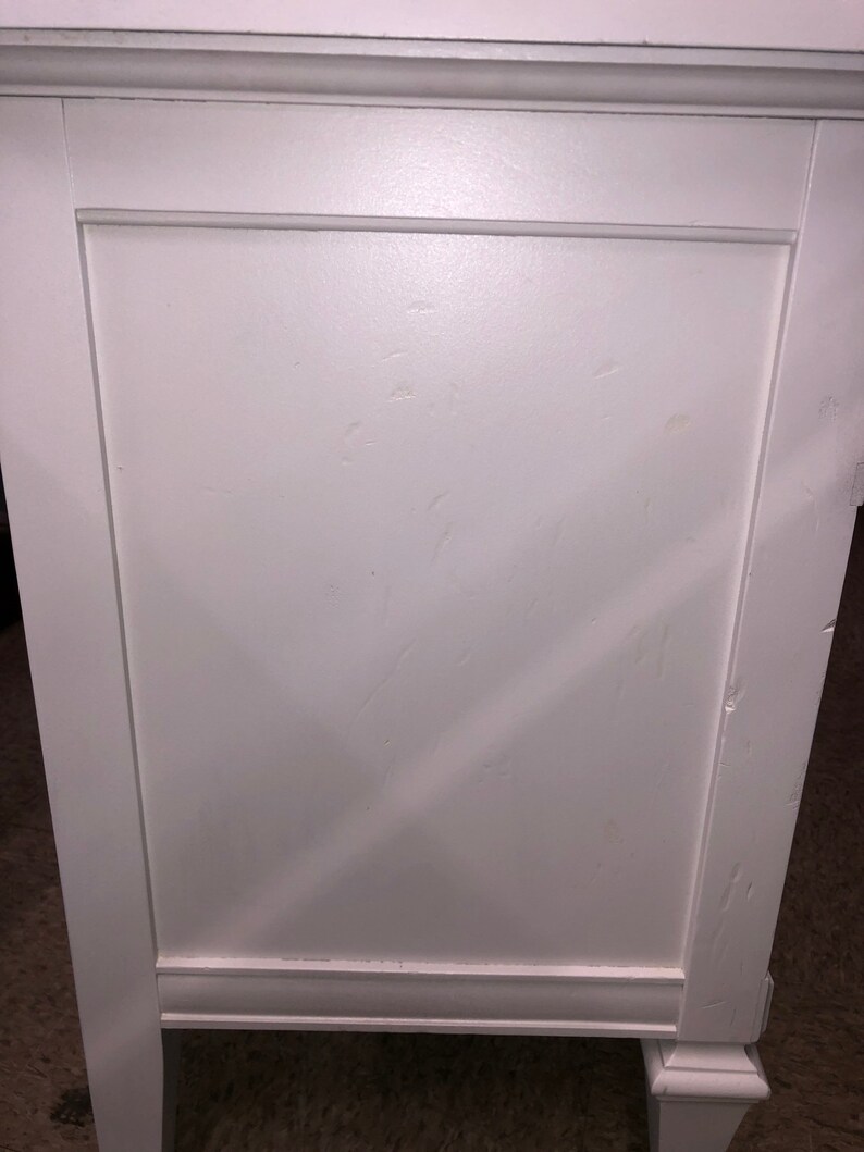 Pu&ograve; includere: Com&ograve; in legno bianco con due cassetti e uno sportello chiuso. Il com&ograve; ha un design semplice con un bordo leggermente rialzato intorno alla parte superiore e inferiore.