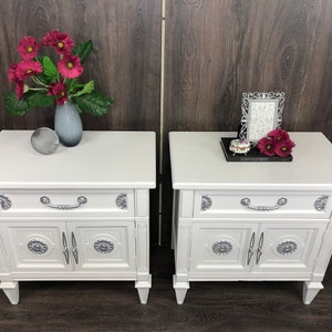 Pu&ograve; includere: Due comodini bianchi con dettagli argentati, ognuno con un cassetto e due ante. I comodini sono su un pavimento di legno. Un comodino ha un vaso con fiori rosa e una sfera argentata. L'altro comodino ha una cornice per foto e una piccola scatola.