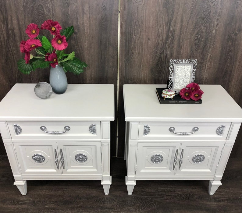 Pu&ograve; includere: Due comodini bianchi con dettagli argentati, ognuno con un cassetto e due ante. I comodini sono su un pavimento di legno. Un comodino ha un vaso con fiori rosa e una sfera argentata. L'altro comodino ha una cornice per foto e una piccola scatola.