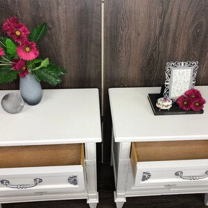 Pu&ograve; includere: Due comodini bianchi con cassetti, uno con un vaso di fiori rosa e una sfera decorativa, l'altro con una cornice, un libro e fiori rosa.