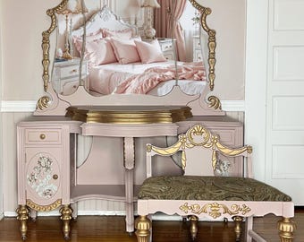 May include: A vintage vanity set with a large ornate mirror, a pink vanity table with gold accents, and a matching pink and gold stool. The vanity table has a drawer and a shelf. The stool has a green paisley patterned cushion.