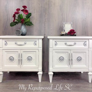 May include: Two white nightstands with silver accents and a drawer and two doors. The nightstands are on a wood floor in front of a gray wood wall.