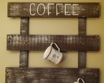 Może przedstawiać: Rusztyczny drewniany stojak na kubki do kawy z napisem "COFFEE" namalowanym na biało na górze. Stojak ma trzy półki i haczyki do zawieszania kubków. Dwa białe kubki z kwiatowymi wzorami wiszą na haczykach.