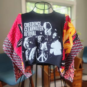 May include: A black cropped sweatshirt featuring a white graphic print of the band Creedence Clearwater Revival. The garment has long, wide sleeves constructed from quilted fabric panels in red, yellow, and black with a checkered pattern.