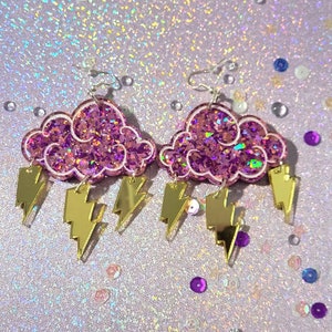 May include: Pink glitter cloud earrings with gold lightning bolt charms. The earrings are made of acrylic and are perfect for adding a touch of whimsy to any outfit.