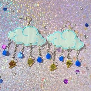 May include: A pair of earrings featuring white cloud charms with blue outlines and silver chains with blue raindrop and gold lightning bolt accents.