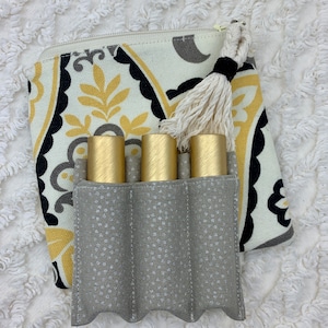 May include: A gray fabric pouch with four slots for holding essential oil roller bottles. The pouch has a white and black paisley pattern and a white tassel.