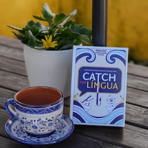 Puede incluir: Un juego de taza y platillo de cerámica azul y blanca con diseños florales se encuentra sobre una mesa de madera junto a una planta en maceta y una caja de juego. La caja del juego dice "Catch the Lingua".