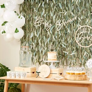 May include: A wooden table with a green leafy backdrop. The table is decorated with a cake, cupcakes, a wooden sign that says "Hey Baby", a glass dispenser, and a wooden crate. The backdrop is decorated with a wooden banner that says "Baby Shower".