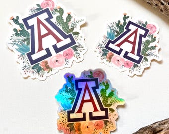 May include: Three stickers featuring the letter "A" in a collegiate font, surrounded by floral and cactus designs. The letter is maroon with a navy border. One sticker has a holographic finish, reflecting multiple colors.