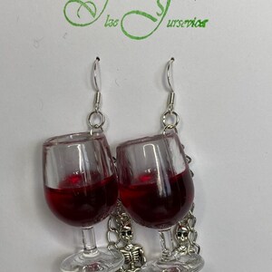 May include: A pair of dangle earrings featuring a glass wine glass filled with red liquid, a silver skeleton charm, and a silver spider charm.