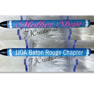 May include: Two blue glitter pens with white accents. The top pen has the text "Mother Shaw" in pink lettering. The bottom pen has the text "JJOA Baton Rouge Chapter". Both pens have the text "TJCreations2" in black lettering.