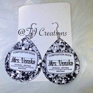 May include: A pair of silver and black glitter teardrop earrings with a white composition book label that reads "Mrs. Vensko" with the details "100 Sheets 200 Pages 9 1/4 in x 7 1/2 in (24.7 cm x 19 cm) Wide Ruled".