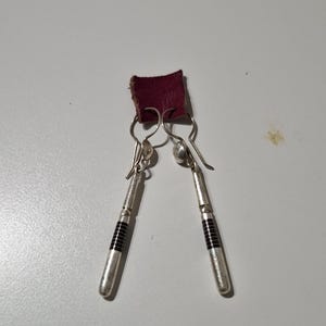 Op de afbeelding: Een paar zilveren oorhangers met zwarte accenten. De oorhangers hebben een kleine zilveren kraal aan de bovenkant en een lange, dunne zilveren hanger met zwarte strepen.