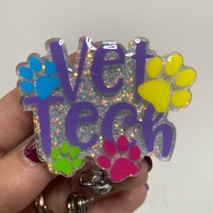 K&ouml;nnte beinhalten: Ein lila glitzernder Badgehalter mit dem Text "Vet Tech" und Pfotenabdr&uuml;cken in Blau, Gelb, Gr&uuml;n und Pink.