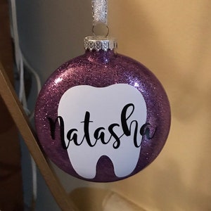 Puede incluir: Adorno de Navidad de purpurina morada con forma de diente blanco y el nombre "Natasha" escrito en negro.
