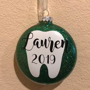 Puede incluir: Adorno de Navidad de color verde con purpurina con forma de diente blanco. El diente tiene el nombre "Lauren" y el a&ntilde;o "2019" escrito en &eacute;l.
