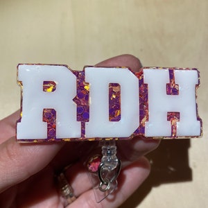 Puede incluir: Portagafete de resina blanca con las letras "RDH" en negrita. Las letras están rellenas de resina brillante de color morado y dorado. El portagafete tiene un carrete de gafete retráctil de plástico transparente.