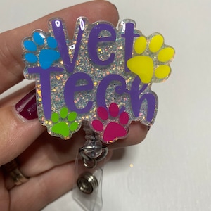K&ouml;nnte beinhalten: Ein glitzernder Badgehalter in Lila mit dem Text "Vet Tech" und bunten Pfotenabdr&uuml;cken. Der Badgehalter hat eine transparente, einziehbare Schnur.