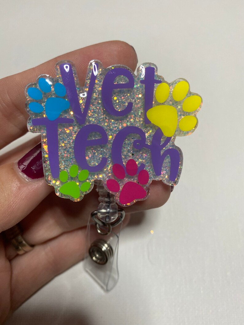K&ouml;nnte beinhalten: Ein glitzernder Badgehalter in Lila mit dem Text "Vet Tech" und bunten Pfotenabdr&uuml;cken. Der Badgehalter hat eine transparente, einziehbare Schnur.