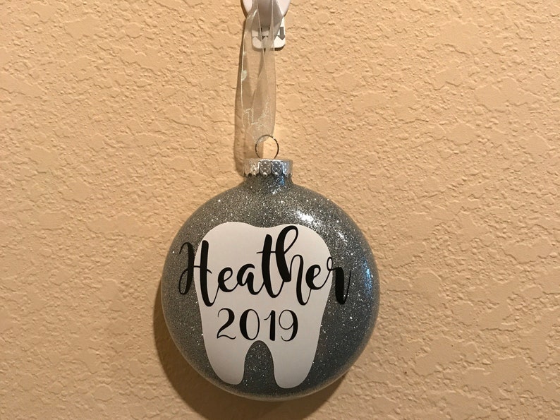 Puede incluir: Un adorno de Navidad de purpurina plateada con forma de diente con el nombre "Heather" y el a&ntilde;o "2019" escrito en negro.