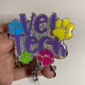 K&ouml;nnte beinhalten: Ein transparenter, glitzernder Badgehalter mit dem Text "Vet Tech" in lila Schrift und bunten Pfotenabdr&uuml;cken. Der Badgehalter hat einen silbernen Drehclip.