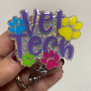 K&ouml;nnte beinhalten: Lilafarbener Glitzer-Badgehalter mit dem Text "Vet Tech" und Pfotenabdr&uuml;cken in Blau, Gelb, Gr&uuml;n und Pink.