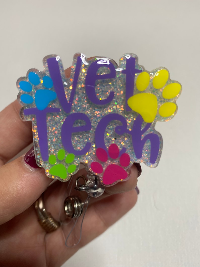 K&ouml;nnte beinhalten: Lilafarbener Glitzer-Badgehalter mit dem Text "Vet Tech" und Pfotenabdr&uuml;cken in Blau, Gelb, Gr&uuml;n und Pink.