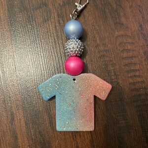 Puede incluir: Un llavero de purpurina azul, rosa y plateado con un dije en forma de camiseta. El dije en forma de camiseta es un degradado de azul claro y rosa con purpurina.