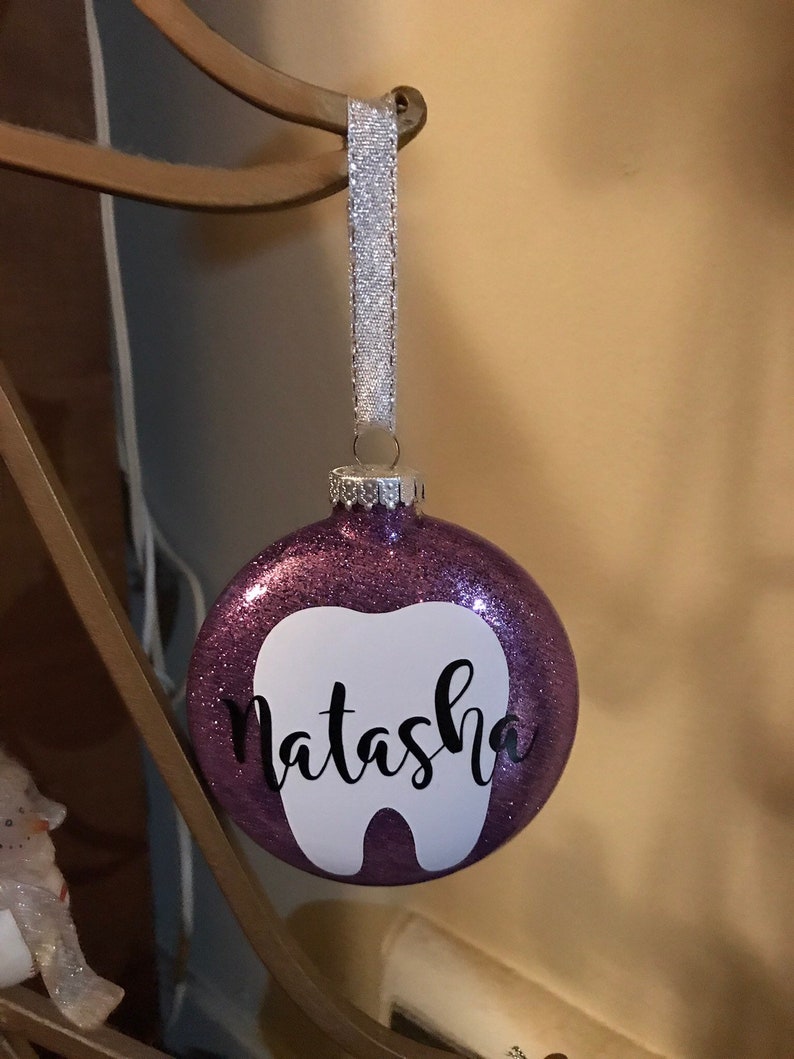 Puede incluir: Adorno de Navidad de purpurina morada con forma de diente blanco y el nombre "Natasha" escrito en negro.