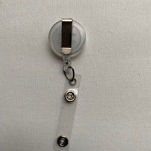 K&ouml;nnte beinhalten: Ein transparenter ausziehbarer Ausweishalter mit einem silbernen Clip und zwei silbernen Ringen. Der Halter ist an einem transparenten Plastikband befestigt.