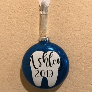 Puede incluir: Adorno navide&ntilde;o azul con purpurina con un dise&ntilde;o de diente blanco y el nombre "Ashley" y el a&ntilde;o "2019" escritos en negro.