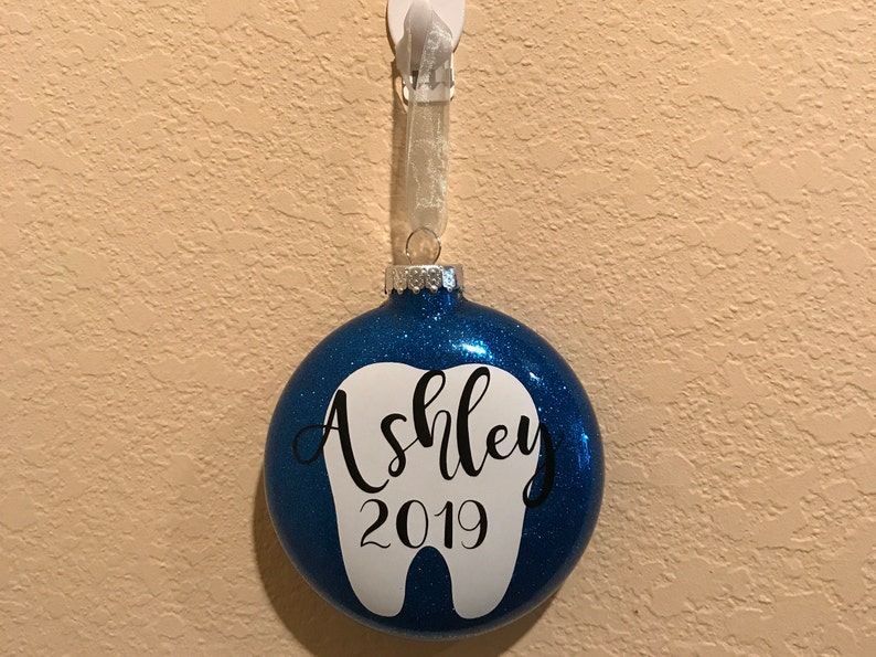 Puede incluir: Adorno navide&ntilde;o azul con purpurina con un dise&ntilde;o de diente blanco y el nombre "Ashley" y el a&ntilde;o "2019" escritos en negro.