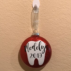 Puede incluir: Adorno de Navidad de color rojo con purpurina con forma de diente blanco y el nombre "Madelyn" y el a&ntilde;o "2019" escritos en &eacute;l.