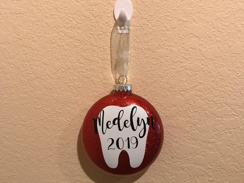 Puede incluir: Adorno de Navidad de color rojo con purpurina con forma de diente blanco y el nombre "Madelyn" y el a&ntilde;o "2019" escritos en &eacute;l.