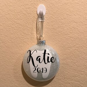 Puede incluir: Un adorno de Navidad de purpurina plateada con un diente blanco y el nombre "Katie" y el a&ntilde;o "2019" escritos en negro.
