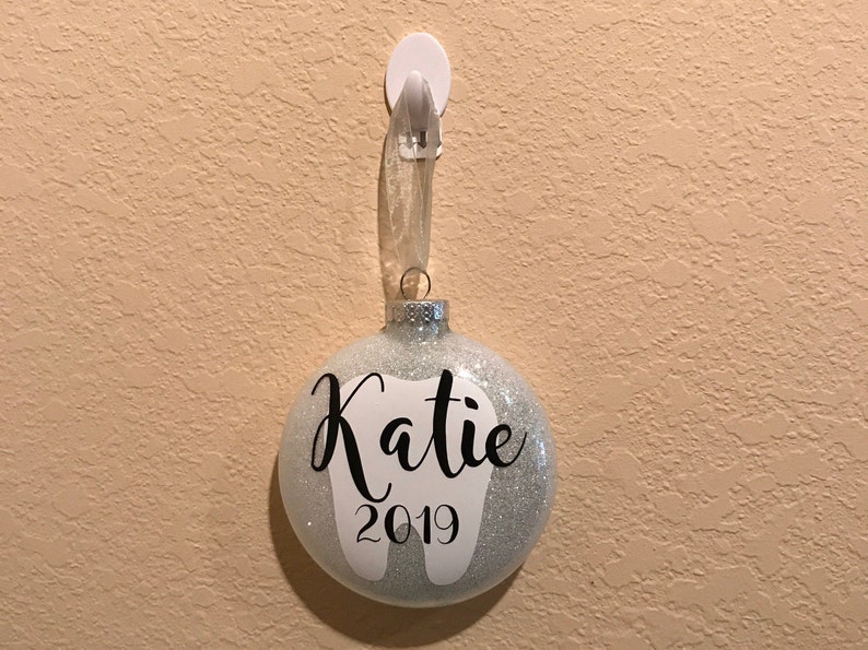 Puede incluir: Un adorno de Navidad de purpurina plateada con un diente blanco y el nombre "Katie" y el a&ntilde;o "2019" escritos en negro.