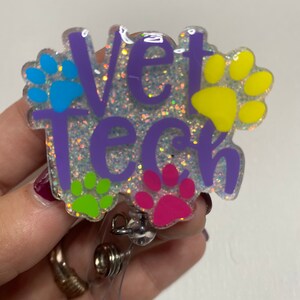 K&ouml;nnte beinhalten: Ein lila glitzernder Badgehalter mit dem Text "Vet Tech" und Pfotenabdr&uuml;cken in Blau, Gr&uuml;n, Pink und Gelb.