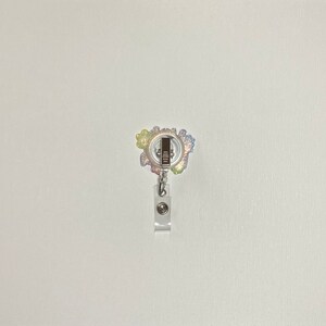 K&ouml;nnte beinhalten: Ein farbenfroher Glitzer-Badge-Rollenhalter mit einem silbernen Clip und einem durchsichtigen Plastikband. Der Rollenhalter ist mit einem Regenbogen aus Glitzer verziert und hat einen silbernen Metallring in der Mitte.