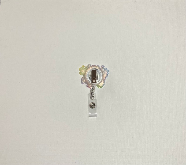 K&ouml;nnte beinhalten: Ein farbenfroher Glitzer-Badge-Rollenhalter mit einem silbernen Clip und einem durchsichtigen Plastikband. Der Rollenhalter ist mit einem Regenbogen aus Glitzer verziert und hat einen silbernen Metallring in der Mitte.