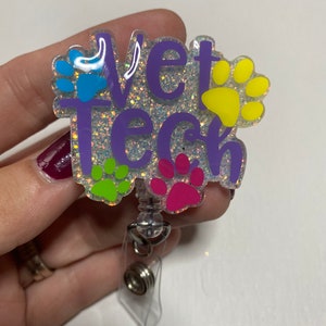 K&ouml;nnte beinhalten: Ein lila glitzernder Badgehalter mit dem Text "Vet Tech" und Pfotenabdr&uuml;cken in Blau, Gr&uuml;n, Pink und Gelb.