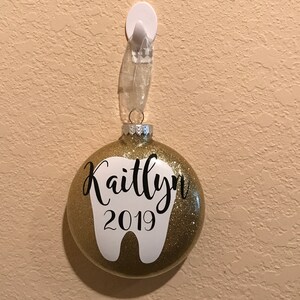 Puede incluir: Adorno navide&ntilde;o de purpurina dorada con forma de diente blanco y el nombre Kaitlyn y el a&ntilde;o 2019.